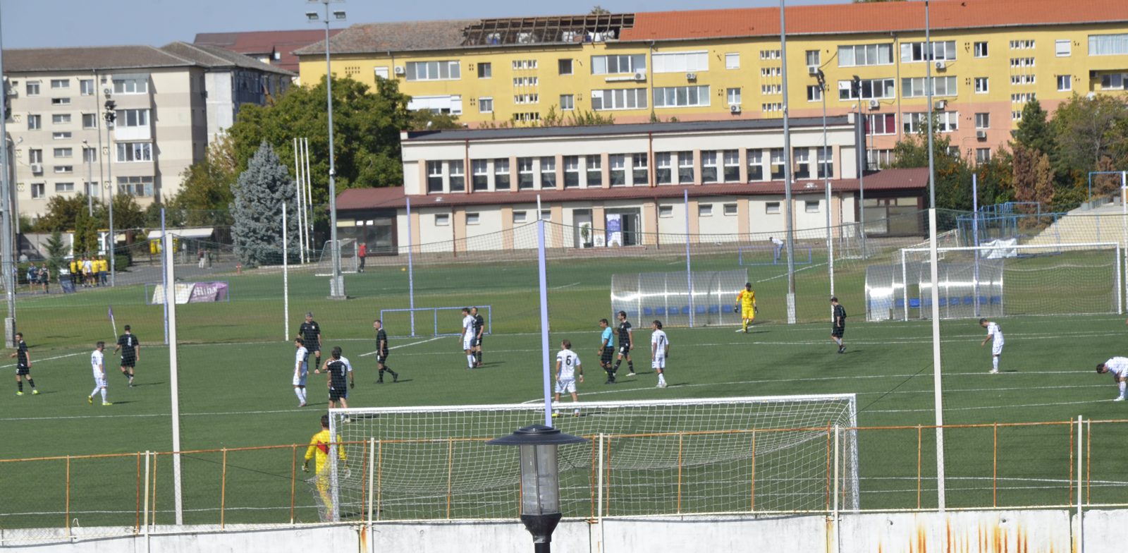 Meci de fotbal între jucători în alb și jucători în negru pe un teren sintetic, cu blocuri în fundal