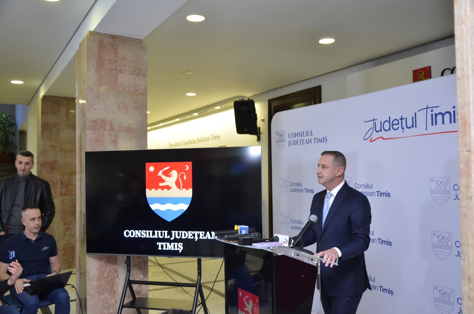 Alfred Simonis la pupitru, lângă ecranul cu stema Consiliului Județean
