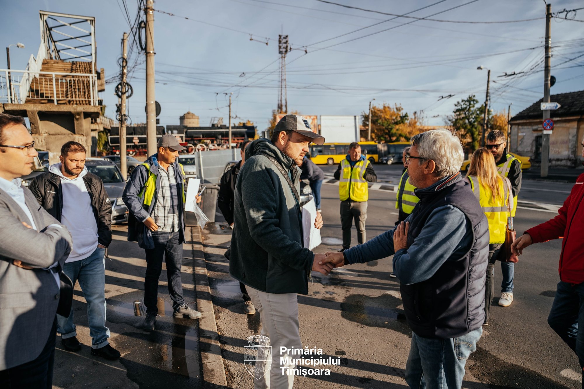 Doi bărbați își strâng mâna, în fața unui grup de persoane în veste reflectorizante