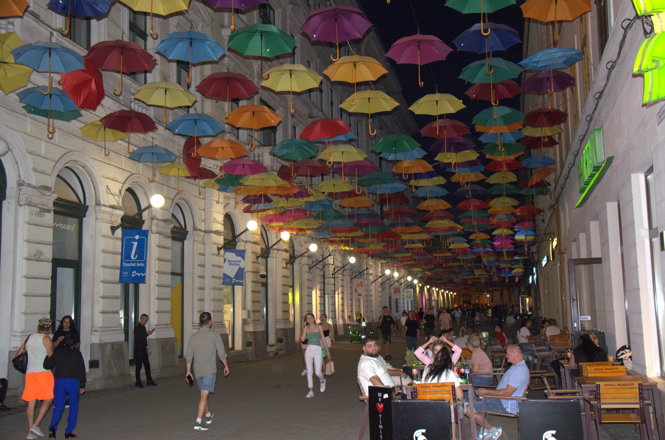 Strada pietonală cu umbrele colorate suspendate, seara, plină de oameni
